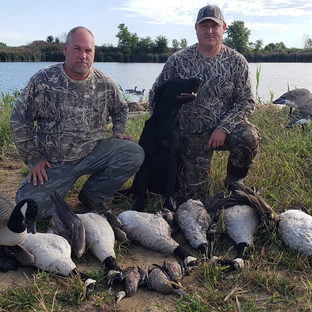 two hunters and a dog kneeling behind a row of geese and game birds